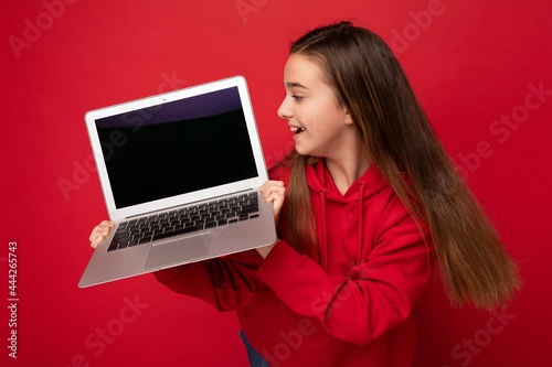 Side profile photo of beautiful happy girl with long hair wearing red hoodie holding computer laptop looking at netbook keyboard and screen monitor isolated over red wall