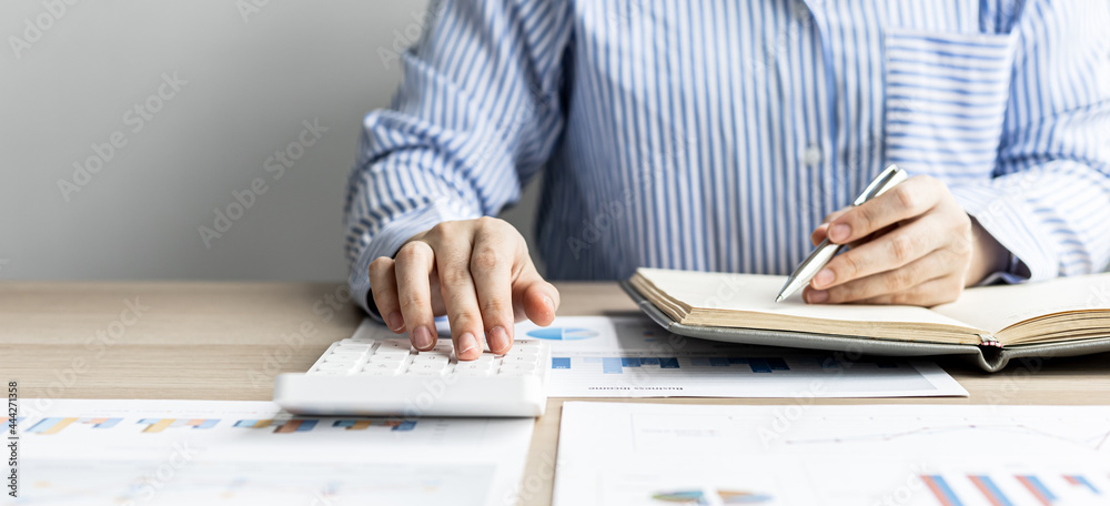 A businesswoman presses on a white calculator and looks at the numbers from a notebook she notes during a finance department meeting to check the accuracy of the numbers on the report. Finance concept A businesswoman presses on a white calculator and looks at the numbers from a notebook she notes during a finance department meeting to check the accuracy of the numbers on the report. Finance concept