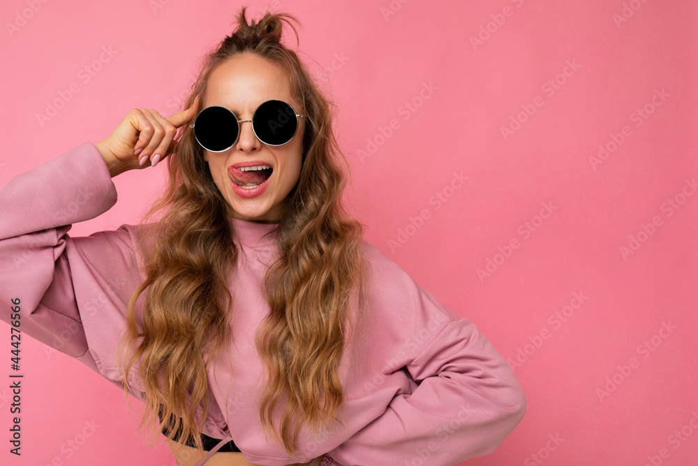 Shot of charming sexy happy young dark blonde curly woman isolated over pink background wall wearing casual pink sport clothes and stylish sunglasses looking at camera and showing tongue Shot of charming sexy happy young dark blonde curly woman isolated over pink background wall wearing casual pink sport clothes and stylish sunglasses looking at camera and showing tongue