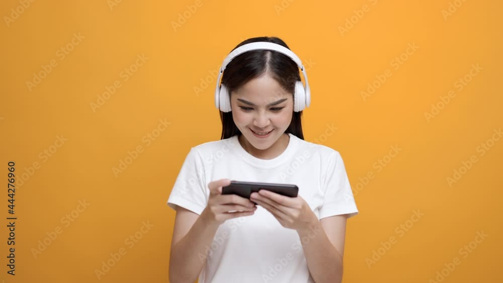 Excited young asian women play mobile game and put on wireless headphone standing on isolated yellow background. Playing game on smartphone winning victory moment. Very enjoy and fun relax time Excited young asian women play mobile game and put on wireless headphone standing on isolated yellow background. Playing game on smartphone winning victory moment. Very enjoy and fun relax time