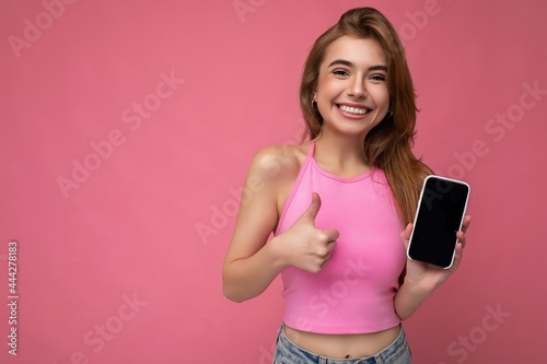 Beautiful joyful young blonde woman wearing pink top poising isolated on pink background with empty space holding in hand and showing mobile phone with empty display for mockup looking at camera and