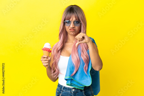 Young mixed race woman with pink hair holding ice cream isolated on yellow background showing thumb down with negative expression