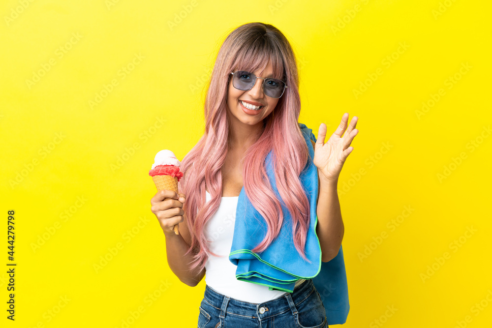 Young mixed race woman with pink hair holding ice cream isolated on yellow background saluting with hand with happy expression Young mixed race woman with pink hair holding ice cream isolated on yellow background saluting with hand with happy expression