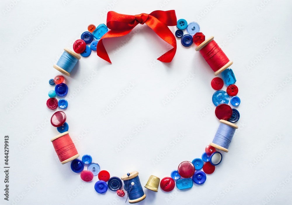 Wreath is made of buttons and threads of red and blue colors with pins and a bright bow of satin ribbon at the top. On a white background with copy space. Concept of craft, sewing and embroidery Wreath is made of buttons and threads of red and blue colors with pins and a bright bow of satin ribbon at the top. On a white background with copy space. Concept of craft, sewing and embroidery