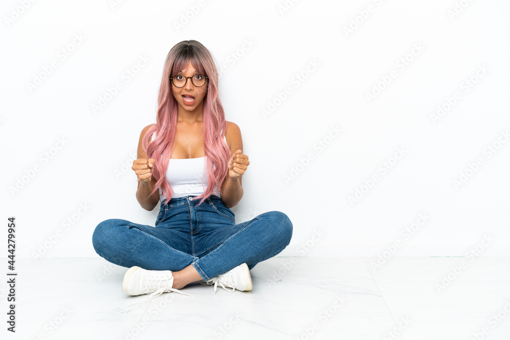 Young mixed race woman with pink hair sitting on the floor isolated on white background celebrating a victory in winner position