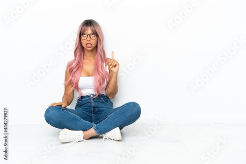Young mixed race woman with pink hair sitting on the floor isolated on white background pointing up and surprised