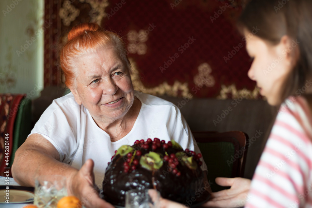 An elderly woman sits at the table and with a smile looks at a young girl,a granddaughter,who wishes her grandmother a happy birthday with a large chocolate cake. Focus on woman. Girl and cake blurred