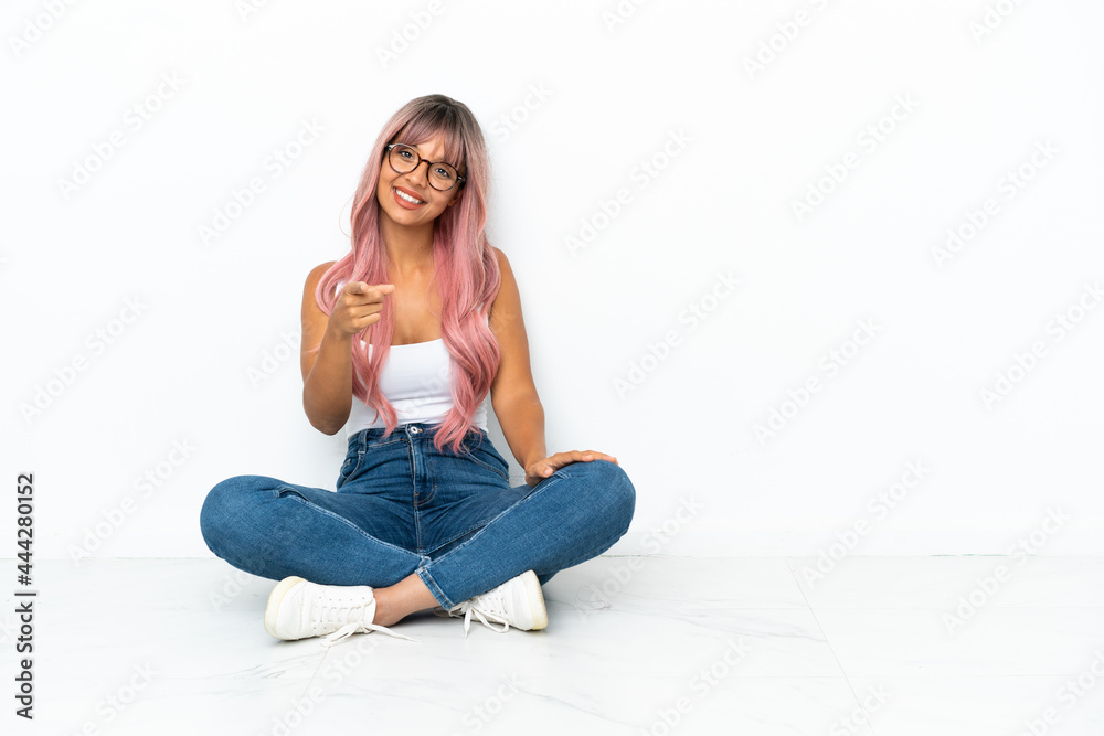 Young mixed race woman with pink hair sitting on the floor isolated on white background points finger at you with a confident expression Young mixed race woman with pink hair sitting on the floor isolated on white background points finger at you with a confident expression