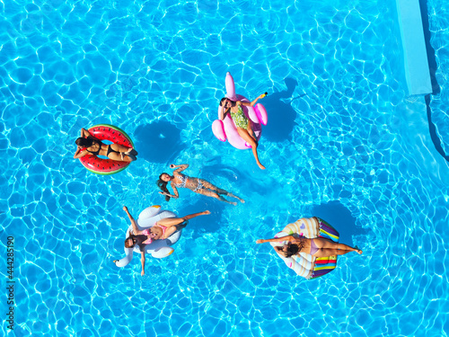 Aerial of hot pretty girls in bikini swimming in pool on floaties. Top view from above. Attractive fitted women in swimsuits relax sunbathing on inflatable flamingo, mattresses. Sunny day summer party