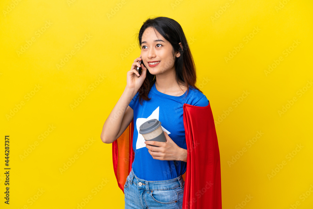 Super hero Vietnamese woman isolated on yellow background holding coffee to take away and a mobile Super hero Vietnamese woman isolated on yellow background holding coffee to take away and a mobile