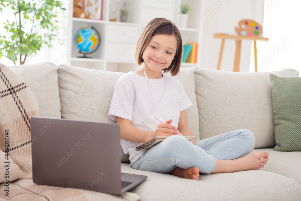 Full size profile side photo of young school girl happy smile listen earphones lesson laptop distance education write notes home Full size profile side photo of young school girl happy smile listen earphones lesson laptop distance education write notes home
