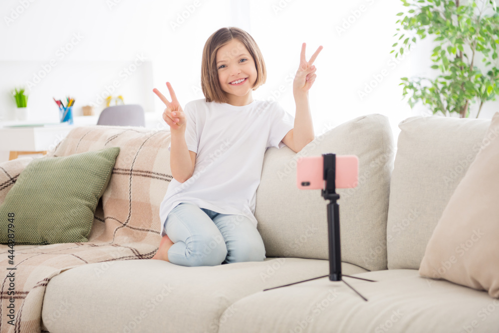 Photo of shiny sweet school girl wear white t-shirt smiling sitting sofa tacking selfie showing v-signs indoors house home room Photo of shiny sweet school girl wear white t-shirt smiling sitting sofa tacking selfie showing v-signs indoors house home room