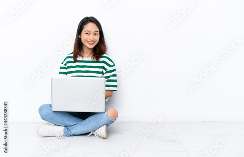 Young Vietnamese woman with a laptop sitting on the floor isolated on white wall with arms crossed and looking forward