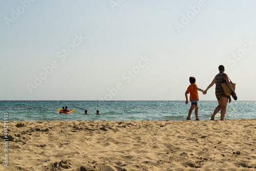Mother and son holding hands on the beach. People in the sea enjoying the beautiful sunny day at Son Bou beach, located in Menorca, Spain. With copy space.