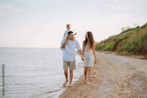 Cheerful young family with little baby boy spending time together on the beach. Father, mother and child against the background of the blue sea and sky. Family, childhood, active lifestyle concept. © maxbelchenko
