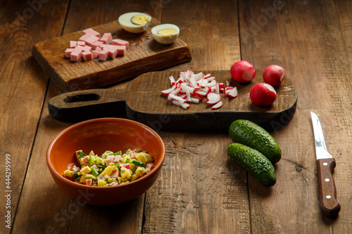 Okroshka in a clay plate on a table on a wooden table next to chopped vegetables and sausage on planks and a knife.