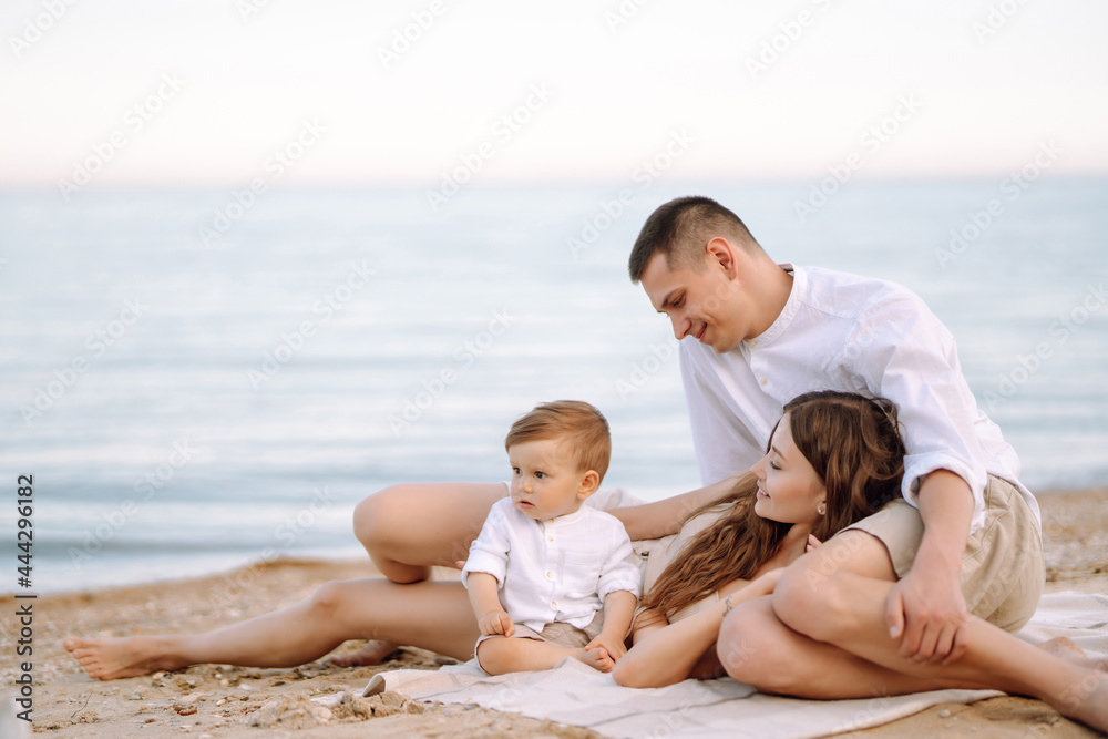 Cheerful young family with little baby boy spending time together on the beach. Father, mother and child against the background of the blue sea and sky. Family, childhood, active lifestyle concept.