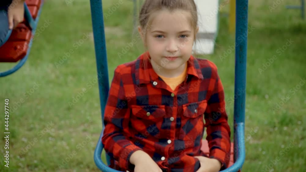 Portrait little girl in red shirt riding on swing. Young happy child with grey eyes on children playard. Inquisitive kid close up. Concept of family holiday and spending time together Portrait little girl in red shirt riding on swing. Young happy child with grey eyes on children playard. Inquisitive kid close up. Concept of family holiday and spending time together