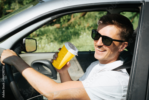 Portrait of a laughing man drinking coffee from a disposable cup while driving a car. Happy handsome driver with glasses, road trip. Summer lifestyle