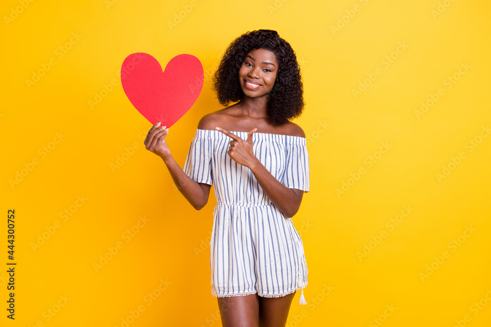 Portrait of beautiful trendy cheerful girl holding in hands demonstrating paper heart card isolated over bright yellow color background Portrait of beautiful trendy cheerful girl holding in hands demonstrating paper heart card isolated over bright yellow color background