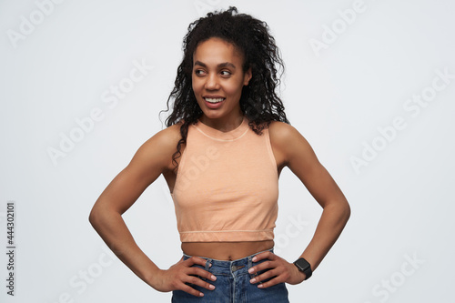 Indoor shot of young african american girl with afro hairstyle, keeps her hand on hips, smiles broadly an looks aside at copy space. isolated over white background
