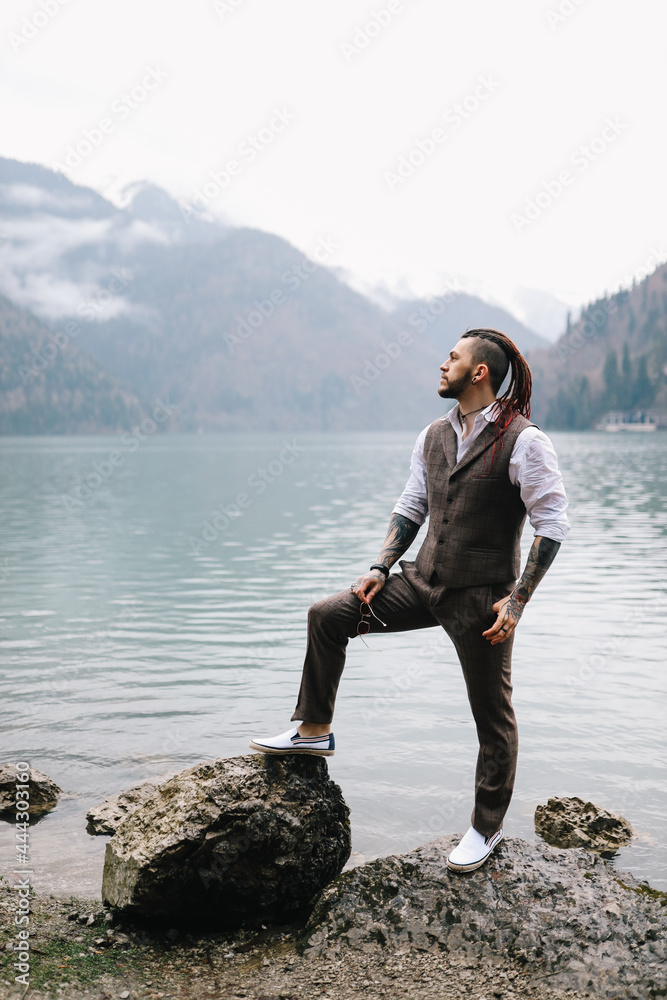 A brutal thoughtful young man groom with a dreadlocked hairstyle in a fashionable elegant wedding suit stands on the shore of a lake against the background of misty mountains in nature