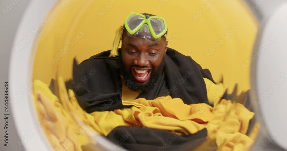 Positive bearded dark skinned man moves as if dancing wears snorkeling mask on forehead poses from inside of washer full of laundry has happy mood poses against yellow background. Washing at home Positive bearded dark skinned man moves as if dancing wears snorkeling mask on forehead poses from inside of washer full of laundry has happy mood poses against yellow background. Washing at home