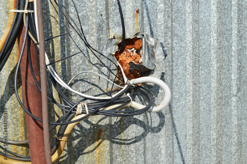 A lot of tangled wires  cables are connected to each other  carelessly come out of a galvanized metal wall.Violation of electrical safety. Electrical wires look dangerous  dirty.