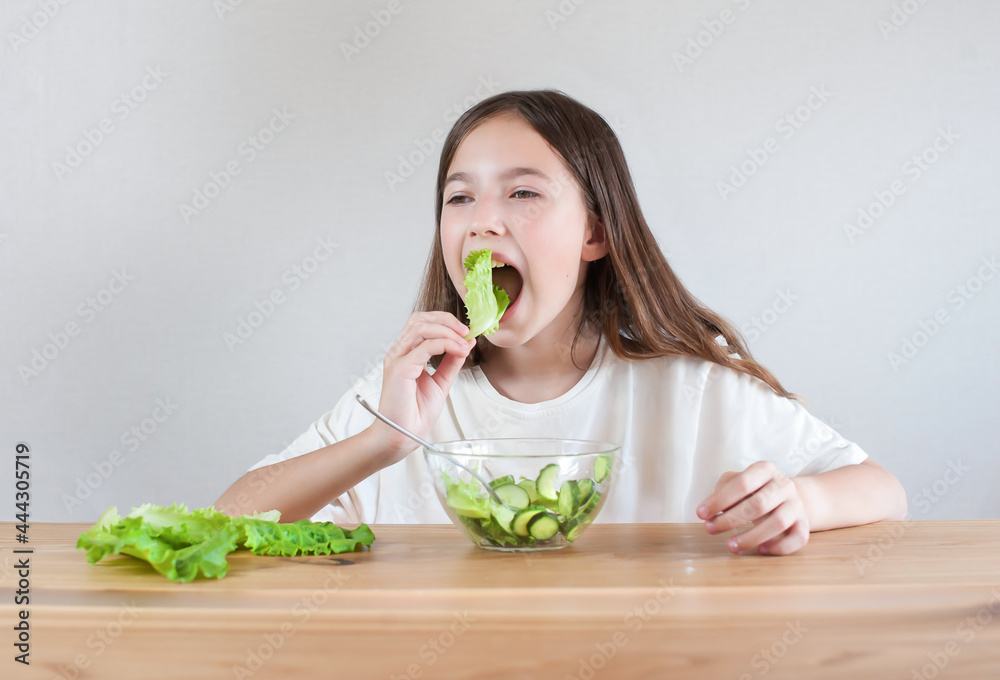 Little girl with long hair having fun eating green salad leaves, vegan vegetable salad sitting at the table Little girl with long hair having fun eating green salad leaves, vegan vegetable salad sitting at the table