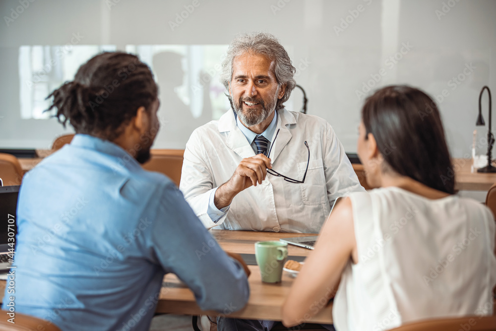 Obraz premium Male doctor consulting with young couple in doctor's room. Male doctor explaining diagnosis to a couple during a visit. Smiling doctor looking at happy couple in medical office