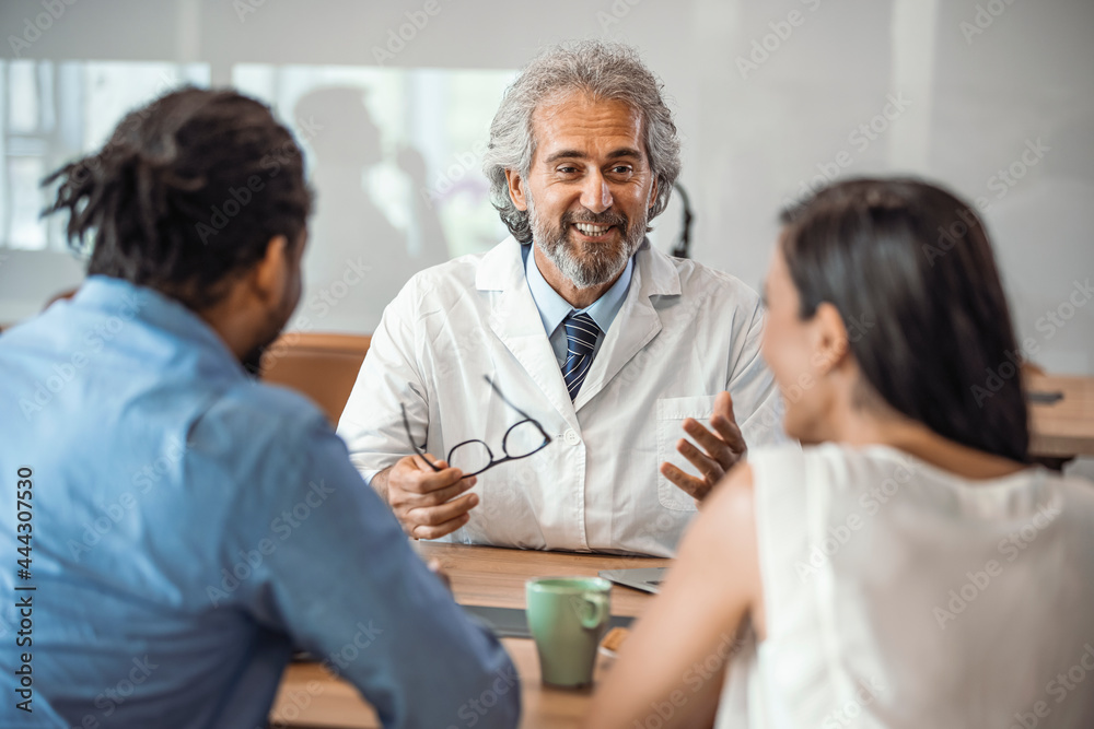 Obraz premium Male doctor offering medical advices to a young couple in office. Husband and wife in a clinic, Beautiful young couple at pregnancy consultation period. Couple meeting medical specialist at hospital