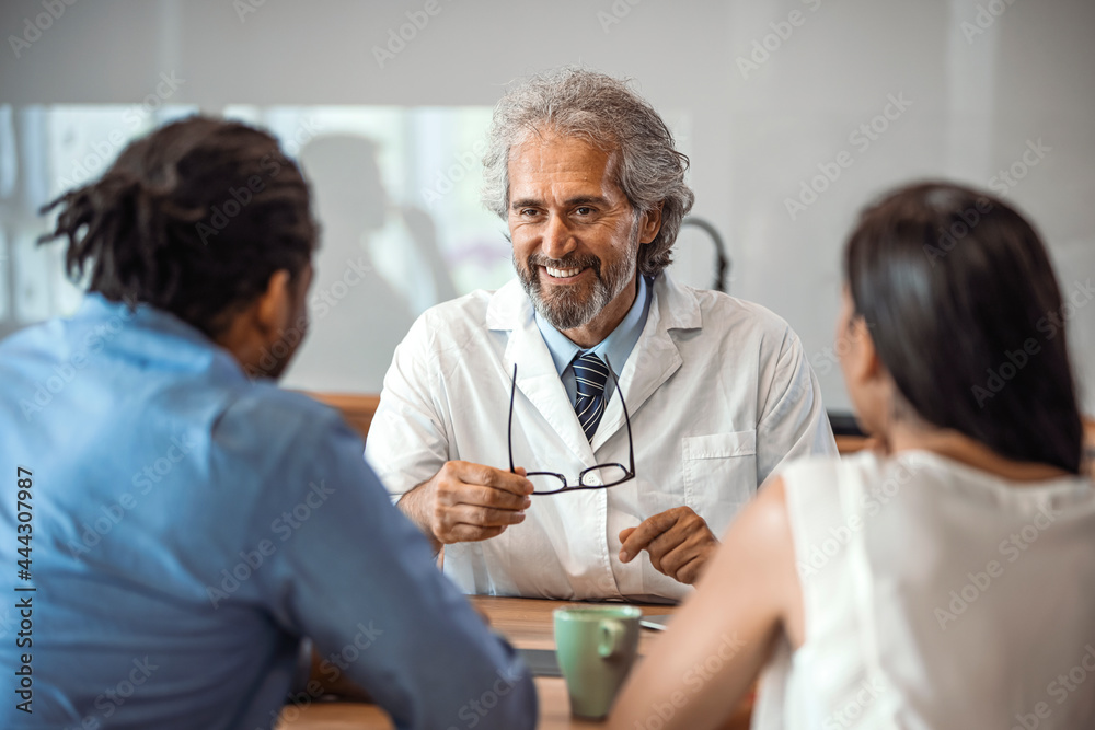 Doctor speaking to young couple. Doctor Using Tablet Computer Discussing Treatment With Patients. Mature Doctor Involved In Serious Discussion With His Patients Doctor speaking to young couple. Doctor Using Tablet Computer Discussing Treatment With Patients. Mature Doctor Involved In Serious Discussion With His Patients