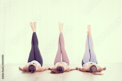 Group of three young sporty attractive women in yoga studio, lying on the floor holdings legs up against white background. Healthy active lifestyle, working out indoors in gym.