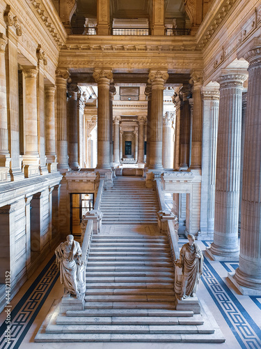 Entrance of the Palais de Justice in Brussels, Belgium. Seeing huge stone pillars surrounding the entrance stairs with greek like statues on a bright day. A marble floor can be seen. 