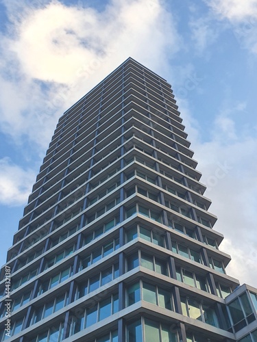 Tall apartment building with glass windows facing a bright blue sky with white clouds in the background. 