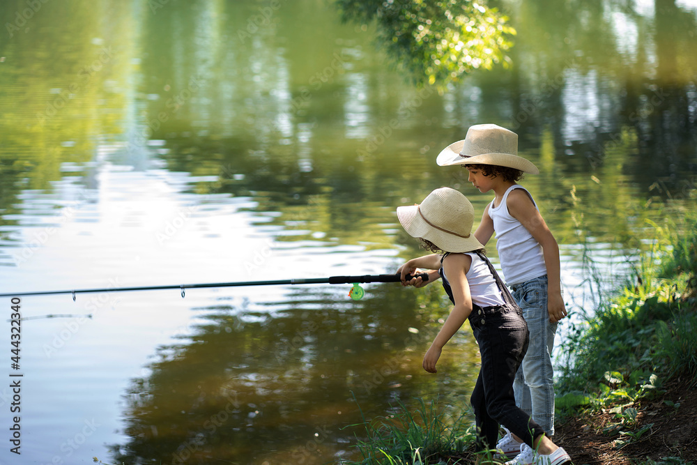 A boy and a girl with curly hair in straw hats stand with a fishing rod by the pond. Children fishing on a summer day. A boy and a girl with curly hair in straw hats stand with a fishing rod by the pond. Children fishing on a summer day.