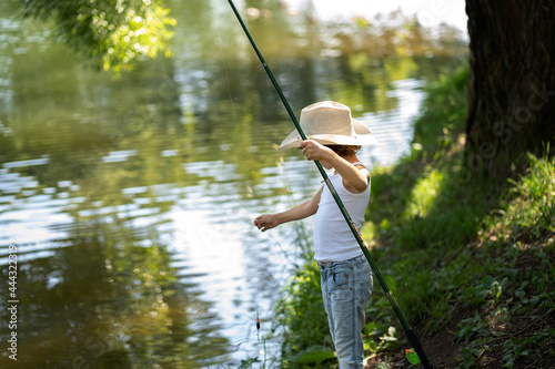 A boy with curly hair in a straw hat stands with a fishing rod by the pond. Child fishing on a summer day.