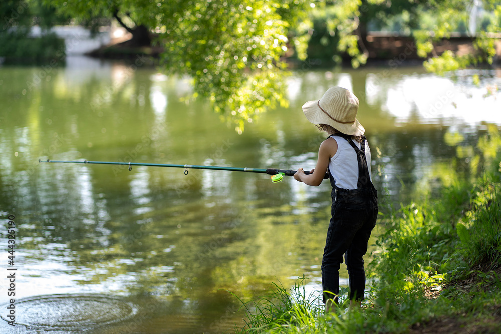 A little girl with curly hair in a straw hat and black overalls stands with  a fishing rod by the pond. Child fishing on a summer day Photos