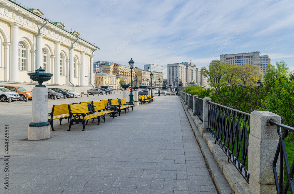 Moscow street with yellow benches and cars parked on it. On the right is a black metal fence.