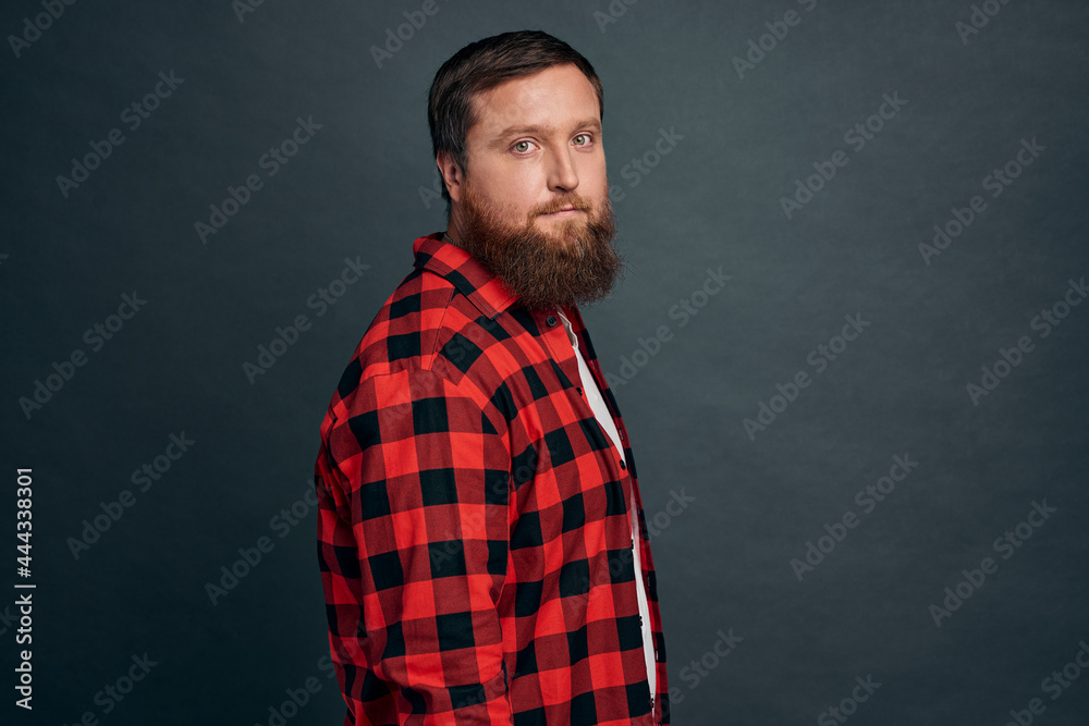 Good-looking modern hipster guy with beard in red checkered shirt, standing half-turned with spaced-out casual expression, looking at camera, posing grey background no emotions, urban people concept.