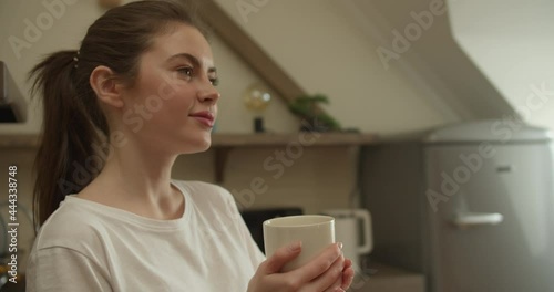 Beautiful brunette caucasian girl in white T-shirt with tail on her head looks away and holds a white cup, then takes one sip and enjoys a drink in the kitchen