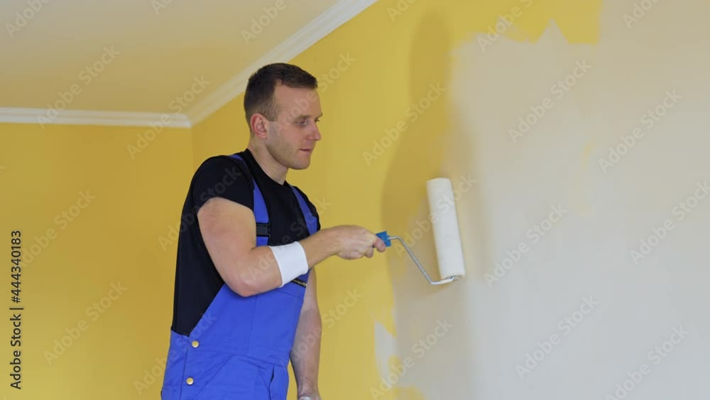 Professional painter paints the walls with roller brush. Worker man in blue overalls renovating and improving flat. Repair and decorating. Professional painter paints the walls with roller brush. Worker man in blue overalls renovating and improving flat. Repair and decorating.