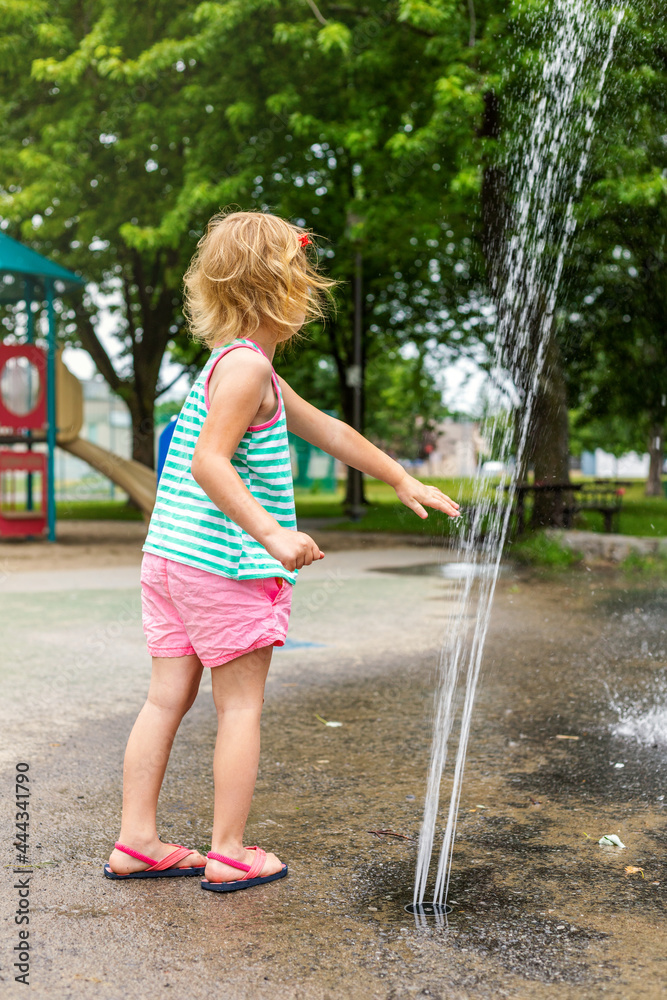 Little happy kid playing with water at splash pad in the local public park on hot summer day. Small cute girl having fun at fountain playground.