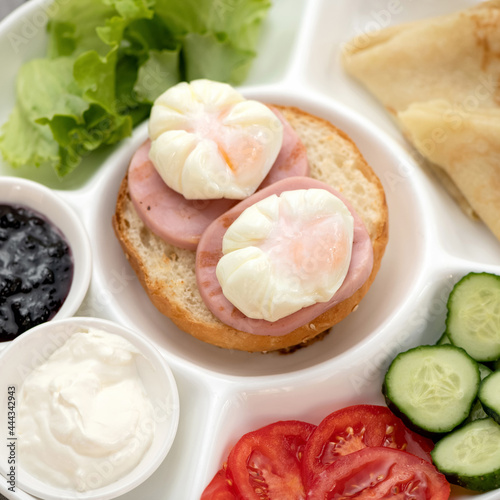 Sandwich with sausage and poached eggs on white portioned plate with vegetables and sauce. Classic European or American Continental breakfast. Close-up shot. Soft focus.