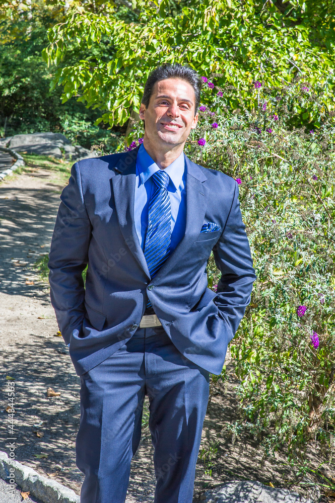 Dressing in a blue suit, blue shirt and blue pattern tie, two hands putting in pockets, a middle age businessman is smilingly standing outside and waiting for you. . Dressing in a blue suit, blue shirt and blue pattern tie, two hands putting in pockets, a middle age businessman is smilingly standing outside and waiting for you. .