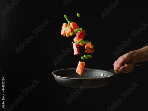 The cook holds in his hand a frying pan with pieces of red fish and vegetables. Levitation. Black background. Contrast. Minimalism. Restaurant dish. Home kitchen.