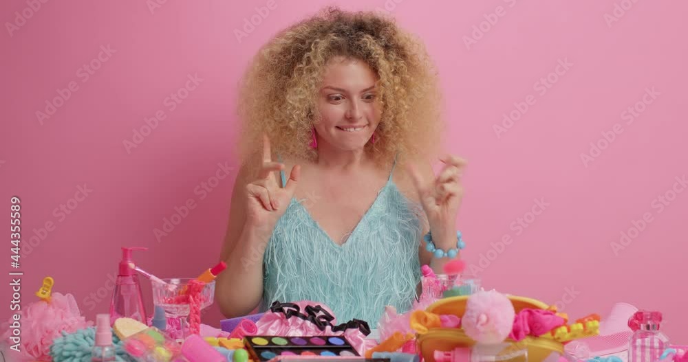 Joyful curly haired young woman looks with surprised happy expression at different cosmetic and beauty products on table wears stylish blue dress going to do makeup isolated on pink background Joyful curly haired young woman looks with surprised happy expression at different cosmetic and beauty products on table wears stylish blue dress going to do makeup isolated on pink background