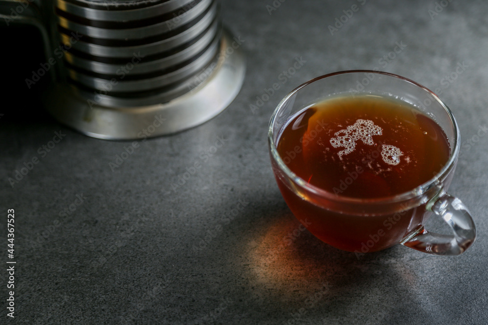 A cup of hot black tea in a glass cup and a kettle press on a gray ceramic countertop. A cup of hot black tea in a glass cup and a kettle press on a gray ceramic countertop.