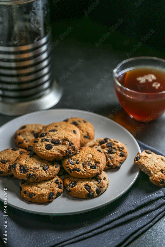 A cup of hot black tea in a glass cup and a teapot press and a gray ceramic plate with cookies with chocolate chips on a gray ceramic counter top with a gray linen napkin.