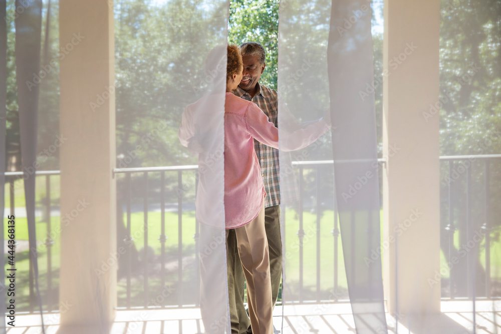 Older couple dancing on porch overlooking green lawn , seen through sheer curtains 